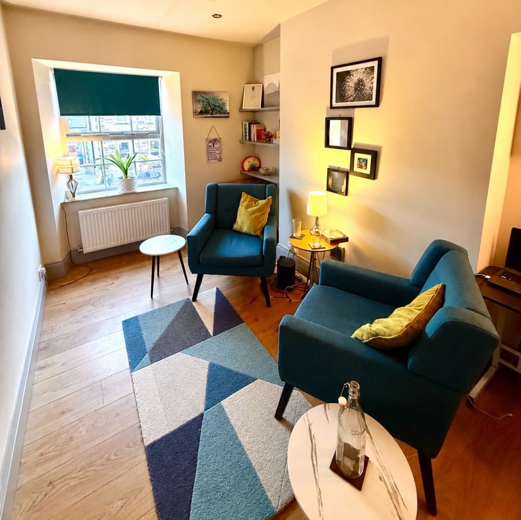 A counselling room with two teal armchairs, mustard and green cushions, a blue geometric rug, round side tables, lamps, a plant beside a window with a dark green blind, and framed pictures on the wall.